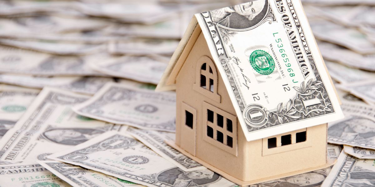 Small model house with a dollar bill placed on the roof, surrounded by scattered dollar bills.